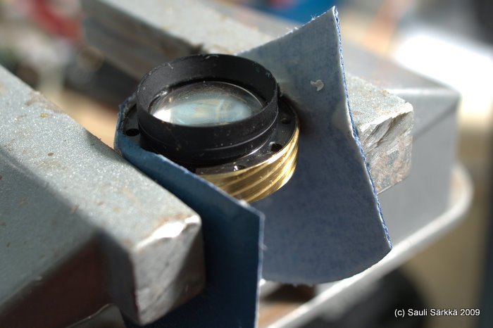 oly_35_rc_front_lens_6.jpg - The elements are encased in a metal tube and the assembly must be secured with just enough force to keep it in place while unscrewing. The blue material is PVC vinyl leatherette that provides cushioning, protection and grip between the helicoid and small bench vise. Mine was fairly tight, but all that I needed to twist off the "cap" was another piece of leatherette...